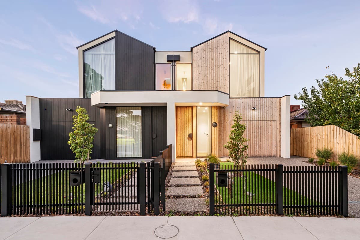 Twin-gabled facade of the Raeburn duplex: black cladding and pale timber battens behind a charcoal picket fence at dusk