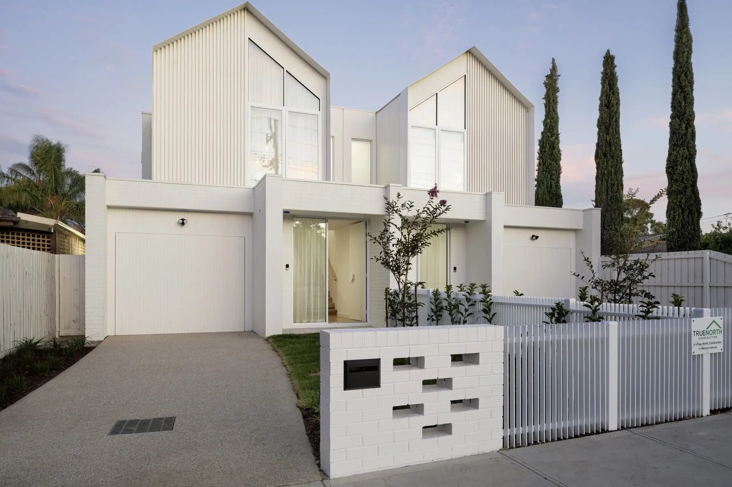 Twin-gabled white facade of the Wingara dual occupancy at dusk, behind a perforated white brick letterbox wall